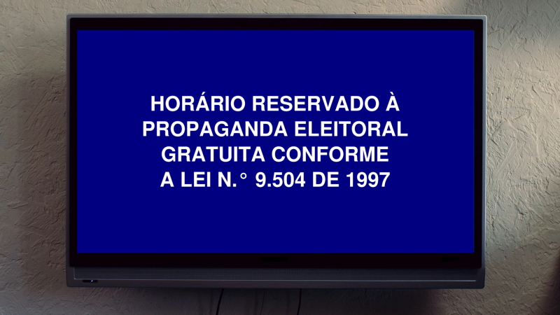 Televisão na parede exibe tela azul onde está escrito: horário reservado à propaganda eleitoral gratuita, conforme a lei número 9504 de 1997.