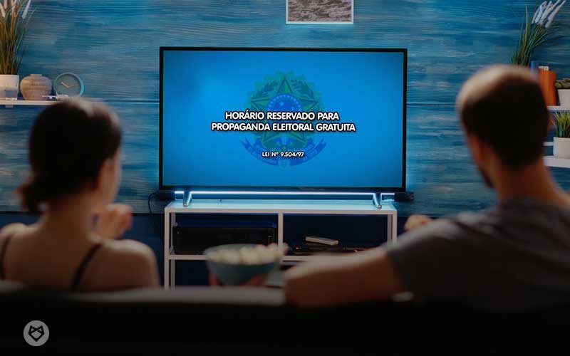 Fotografia mostra casal numa sala, em frente à televisão, assistindo à propaganda eleitoral gratuita.