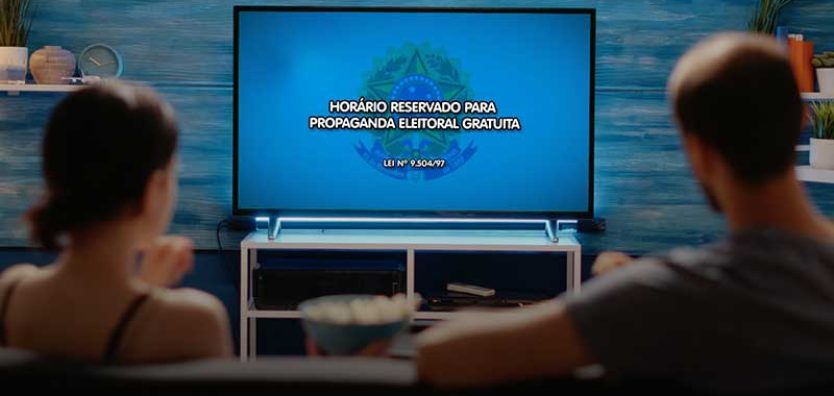 Fotografia mostra casal numa sala, em frente à televisão, assistindo à propaganda eleitoral gratuita.
